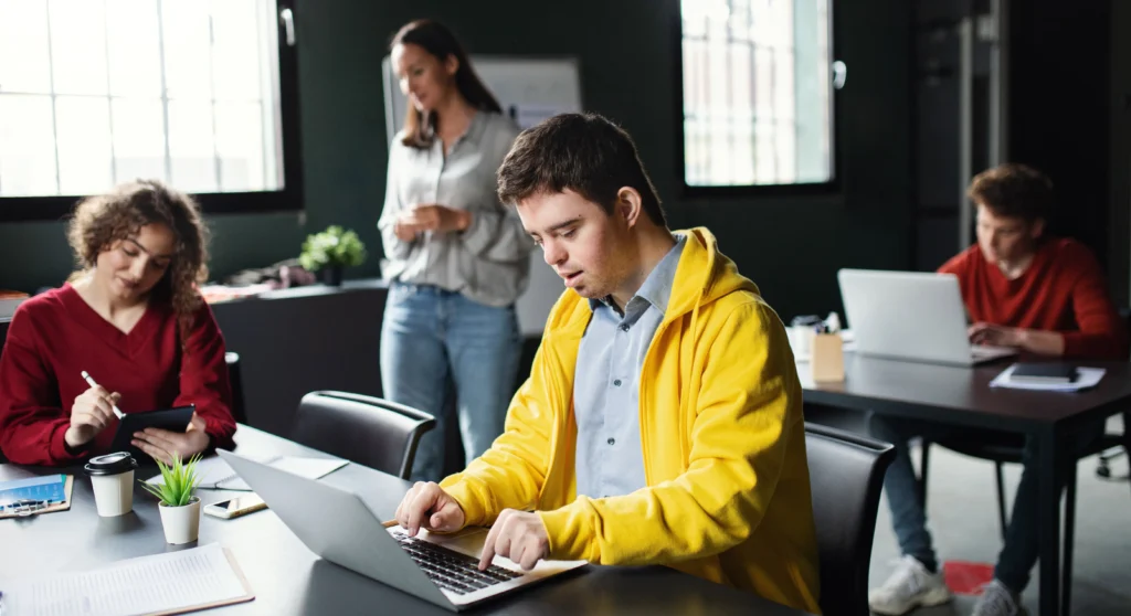Free and Appropriate Public Education. A young disabled man sits at a desk working on a laptop in a classroom. 