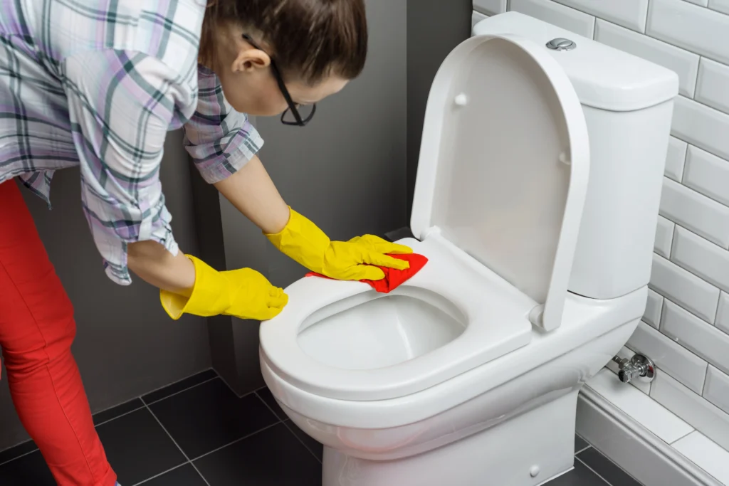 A young female student wearing yellow rubber gloves is leaning the surface of a toilet.